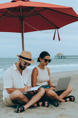 Fototapeta premium Vertical image of Caucasian young adult couple working on beach under umbrella using laptop and writing in notebook, representing remote work lifestyle and digital nomad career concepts