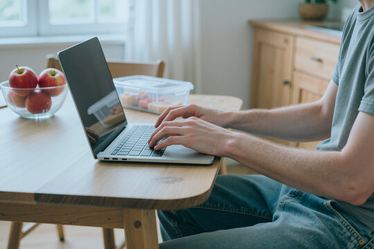 Caucasian young adult man typing on laptop keyboard at kitchen table, working from home. Image represents remote work, online education, and freelance lifestyle, medium shot