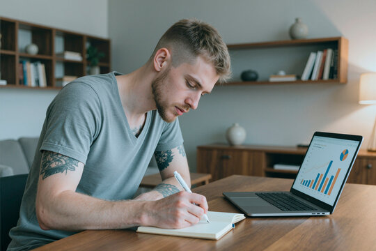 Young adult Caucasian man writing in notebook near laptop with financial charts on screen, working from home office. Image suitable for remote work and freelance business concepts