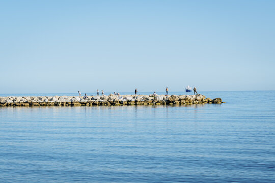 Malaga, Andalusia, Spain - 18 May 2020: View of people standing on a rocky breakwater in the Mediterranean sea under a clear blue sky.