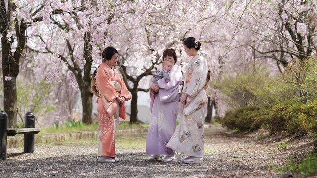 Three adult women in kimono stand together beneath cherry blossoms, with the center woman holding a baby.