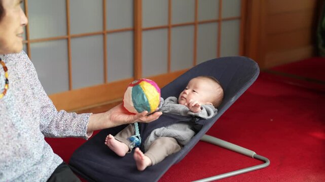 Older woman holds a colorful patchwork ball in front of a baby reclining in a blue bouncer.