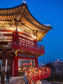 Busan, South Korea - 07 February 2026: View of a traditional Korean pavilion with ornate wooden eaves and red pillars illuminated at dusk, featuring a balcony with string lights.
