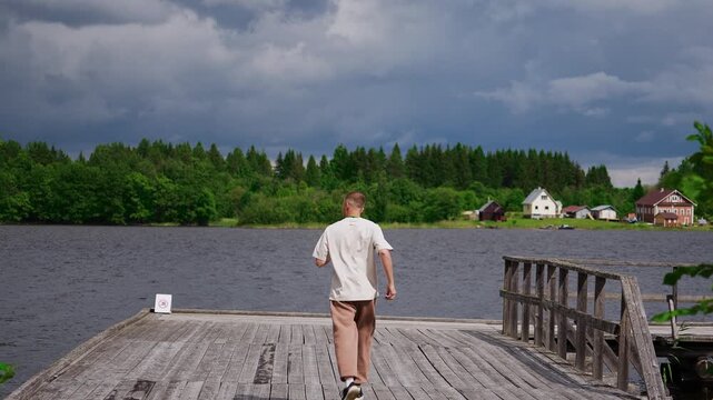 Man running on wooden pier by lake with forest and village on background, Karelia summer
