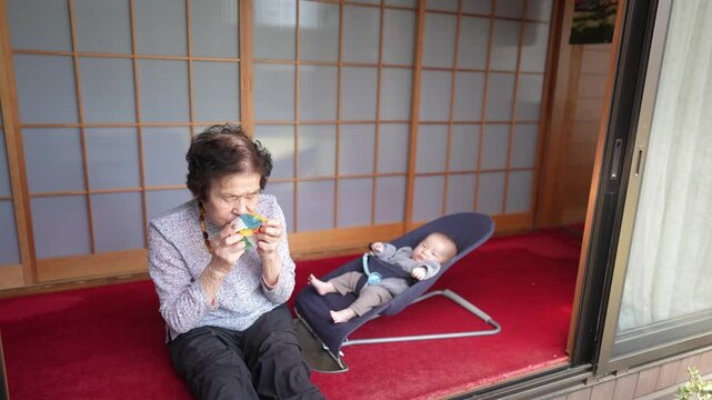 Older woman holds a colorful patchwork ball close to her face while a baby reclines in a bouncer beside her.
