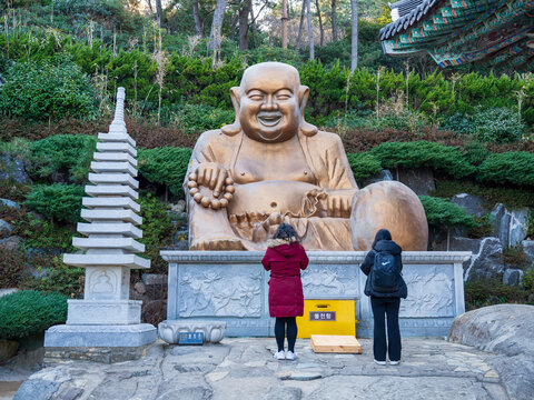 Busan, South Korea - 14 February 2026: View of two people standing before the golden Buddha statue at Haedong Yonggungsa Temple, with a stone pagoda and lush greenery in the background.