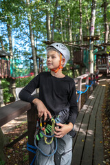 A young boy wearing a safety helmet and climbing harness stands at an outdoor adventure park. He is ready for a rope course activity, representing active lifestyle, courage, and outdoor recreation. © JULIA
