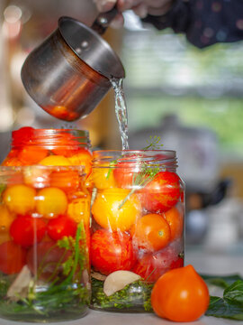 Close-up of glass jars filled with cherry tomatoes, dill and garlic pouring with hot water from saucepan. Traditional homemade food preservation and seasonal harvest canning.