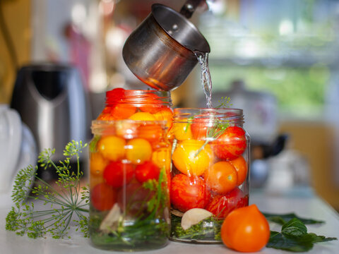 Process of pouring hot water into glass jars filled with fresh tomatoes, dill, garlic for home canning and pickling. Home food preservation, seasonal harvest, preparation for winter.