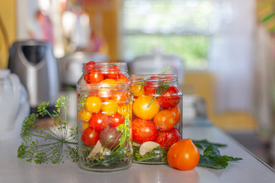 Glass jars filled with fresh colorful cherry tomatoes for pickling in home kitchen. Traditional home food preservation and culinary preparation. Cozy, rustic atmosphere.