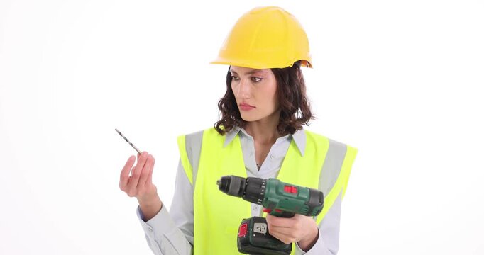 Female builder looks at electric drill and sharp bit with confused expression. Woman in safety helmet and durable work uniform on white background