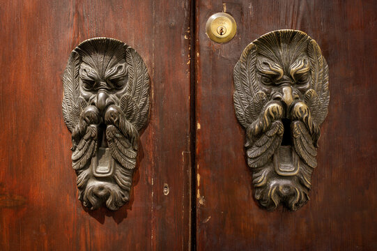 Bologna, Emilia-Romagna, Italy - 17 May 2018: View of ornate bronze door knockers with grotesque faces mounted on a weathered wooden door in the historic city center.