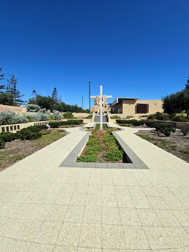 Geraldton, Australia - 29 December 2025: View of a memorial garden featuring a large stone cross, paved walkways, and manicured hedges under a clear blue sky.