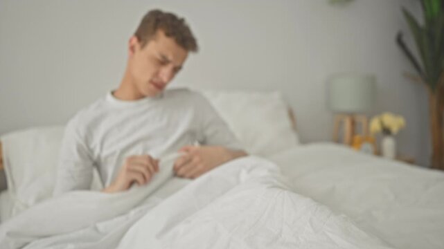 Man rubbing his eyes on a white bed in a building bedroom with lamp, clock, bedside table and potted plant visible, sitting upright under a duvet and pillow; sleepiness.