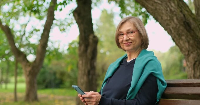 Beautiful smiling mature blond woman with glasses using her phone for texting messages while sitting on bench in the park