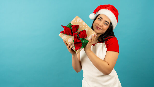 Young latina woman wearing santa hat holding a christmas present on a blue background