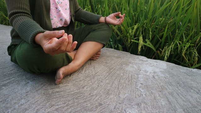 A woman practicing meditation in a peaceful outdoor setting near a green rice field.