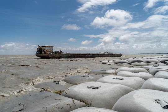 Noakhali, Chittagong Division, Bangladesh - 15 August 2026: View of a weathered barge on turbulent muddy river waters, with large sandbags along the embankment for flood protection under a cloudy sky.