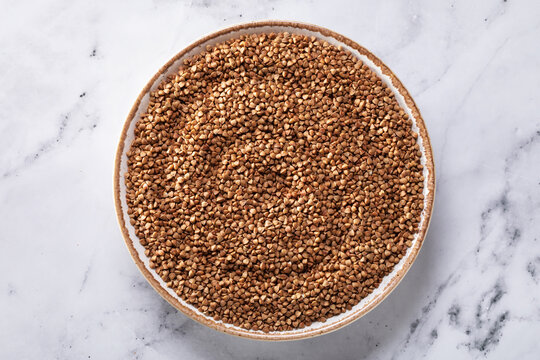 Uncooked buckwheat groats in a ceramic bowl on a marble table. Top view