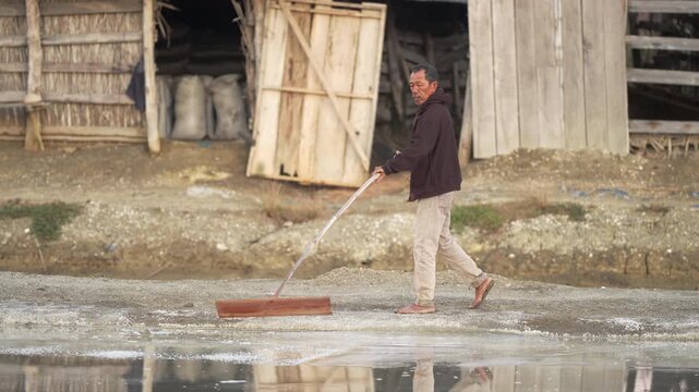 traditional indonesian salt farmer harvesting sea salt with a wooden rake in indonesia, southeast asia, manual worker, slow motion