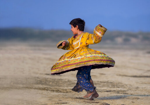 Turbat, Balochistan, Pakistan - 29 November 2017: View of a young child in a vibrant yellow traditional dress spinning and dancing on a sandy landscape.