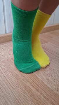 Odd socks day highlights green and yellow socks on bare feet indoors. Mismatched pair shifts position on wood floor, giving casual, cheerful style and clear color contrast during movement.