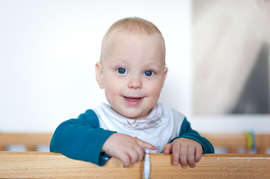 A cheerful baby with bright blue eyes and a playful expression leans over the edge of a wooden crib in a warm, inviting room. The soft light adds to the joyful atmosphere.