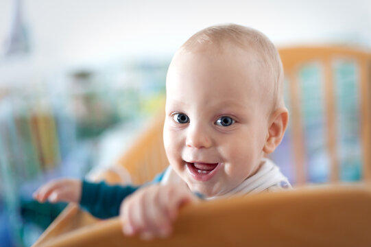 A cheerful baby leans over the wooden crib, flashing a big smile. The soft natural light fills the cozy room, creating a warm and inviting atmosphere.