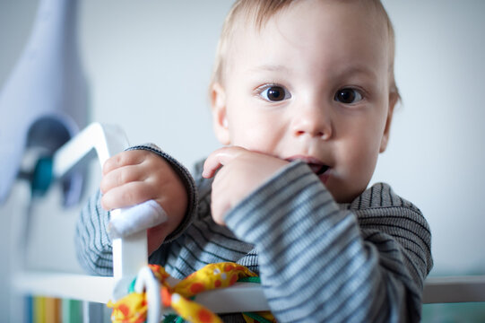 In a cozy home setting, a curious baby leans over the edge of a colorful crib. The child looks inquisitively while chewing on a finger, surrounded by soft toys.