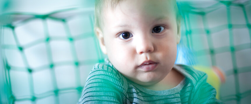 A young child sits in a cozy play area, looking curiously through green netting. The atmosphere is bright and cheerful, filled with colorful toys and warmth, creating a fun environment.
