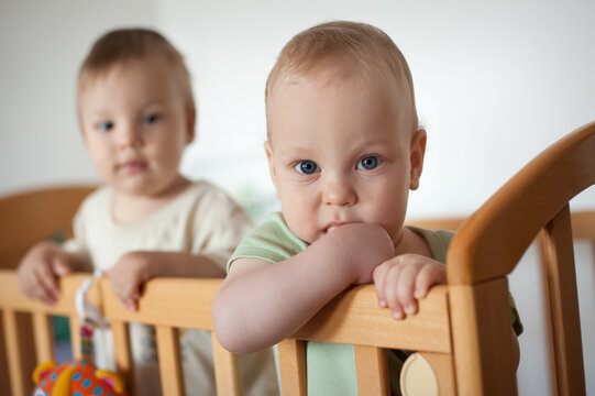 In a bright nursery, two adorable twins are peering over the edge of their wooden crib, discovering their surroundings.