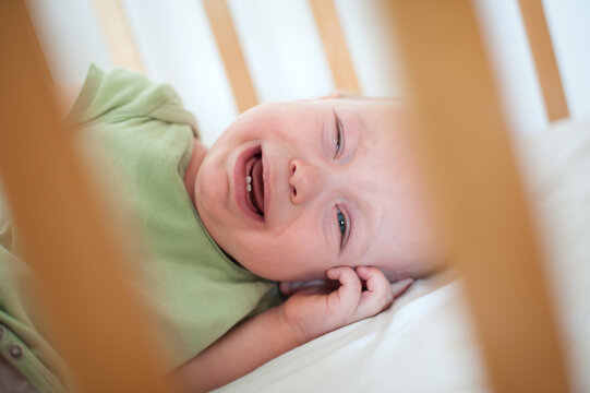 A small baby lies in a wooden crib, tears streaming down their cheeks as they cry. The soft light creates a gentle atmosphere, revealing their distress and need for attention.