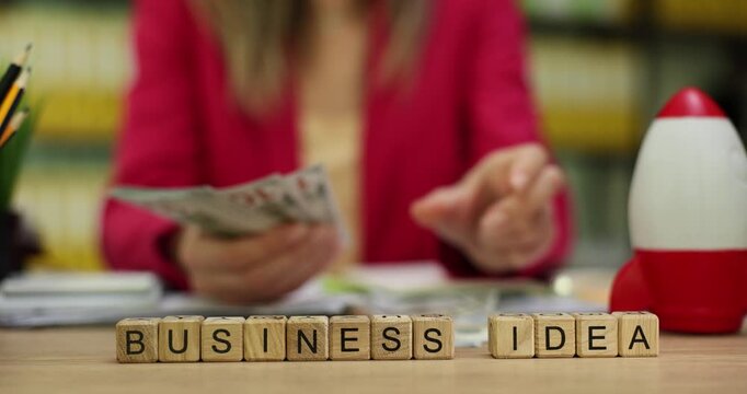 Miniature wooden cubes create phrase Business Idea on desk. Businesswoman holds stack of dollar bills and counts available funds. Slow motion