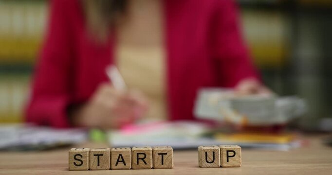 Miniature wooden cubes form phrase Start Up on office table. Businesswoman writes down available financial resources for new project. Slow motion