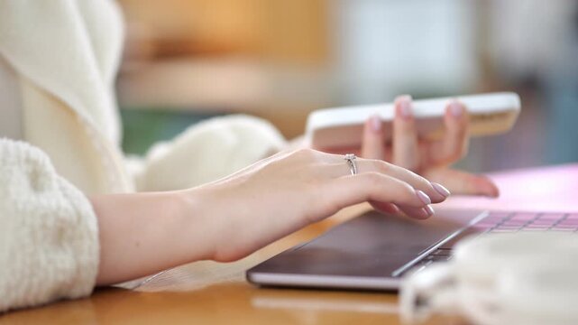 Close-up of hands typing on laptop keyboard. Person wears light sweater with rings on fingers. Fingers move over dark laptop surface. Background softly blurred with warm tones