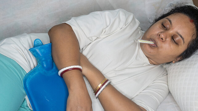 Indian young sick woman checking fever with thermometer during menstrual periods. Using hot water bag to relive cramp or pain, while lying on sofa at home. Fever and illness concept.