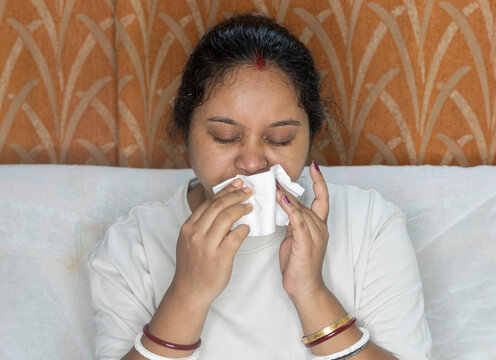Indian young woman sick, suffering from fever and cold flu on sitting sofa, blowing on her nose with napkin or tissue, Pollen allergy, sneezing, flu fever concept.