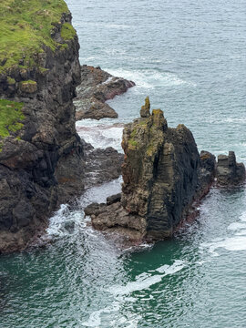 A jagged basalt sea stack rises beside steep volcanic cliffs as Atlantic waves swirl through the rocky shoreline on Northern Ireland&rsquo;s Causeway Coast.