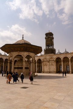 Cairo, Cairo Governorate, Egypt - 19 March 2022: View of the Mosque of Muhammad Ali with its ornate ablution fountain, clock tower, and visitors walking across the stone courtyard under a cloudy sky.