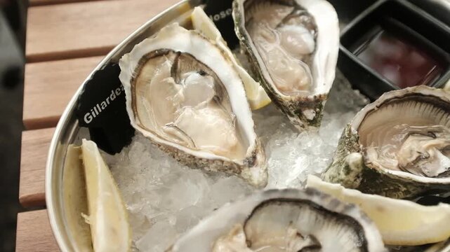 Fresh raw oysters on ice with lemon wedges and a small bowl of sauce in a metal tray. For seafood restaurant promotions, culinary shows, gourmet dining content, or luxury food commercials.