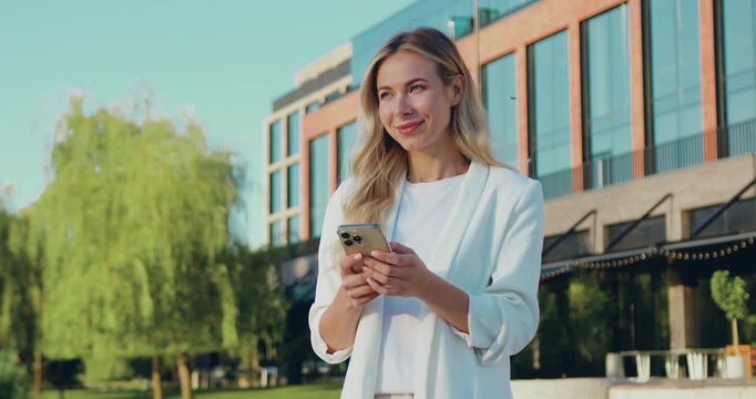 Adult woman wearing a white blazer standing on a city street, using her smartphone to read and reply to text messages.