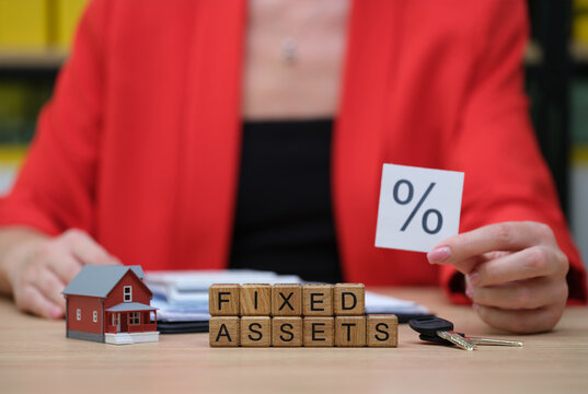In a modern office, a business professional holds a percentage sign while seated at a desk with documents. Nearby, wooden blocks spell out fixed assets and a small house model is present.