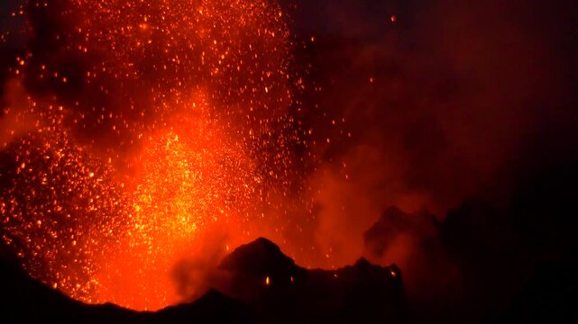View of Stromboli volcano erupting with glowing lava and sparks against the dark night sky in Stromboli, Sicily, Italy.