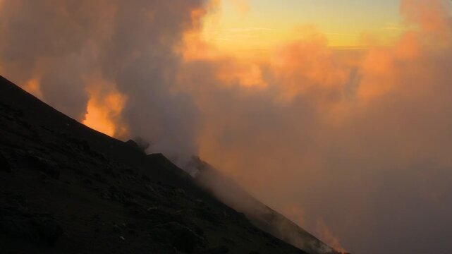 View of Stromboli volcano erupting with thick volcanic smoke and glowing embers against a golden sunset sky in Stromboli, Sicily, Italy.