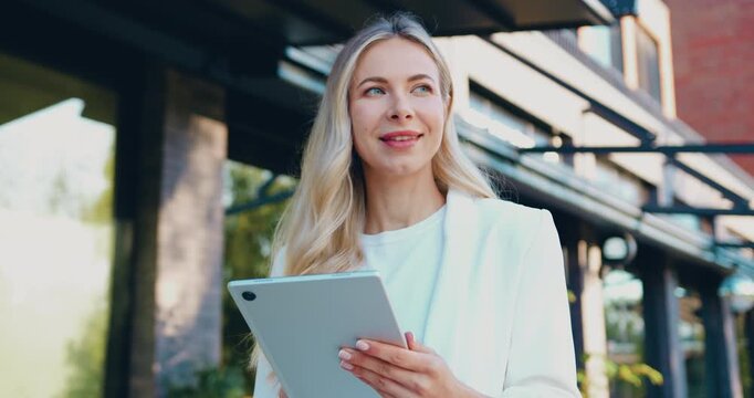 Portrait of a young woman holding a tablet while shopping online outdoors, smiling and looking away, browsing products and making online purchases on a city street