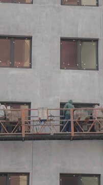 Construction workers finishing and plastering the building facade while standing in a suspended cradle. Operatives are plastering the exterior wall of a multi storey house at high altitude