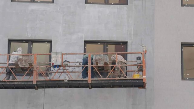 Construction workers performing facade finishing and plastering while standing in a suspended cradle. Operatives plaster the exterior wall of a multi storey building near the windows at high height