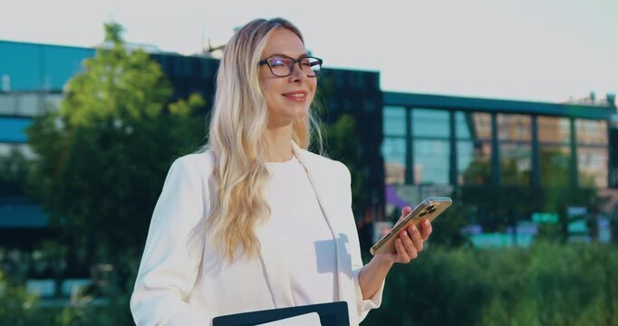 Businesswoman texting on a smartphone while walking near a corporate office building, checking emails and communicating online.