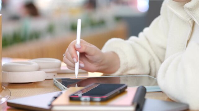 Person uses stylus on tablet. Hands focus on digital drawing. Modern tech tools on wooden desk. Soft lighting highlights creative workspace. Objects include phone, notebook, headphones