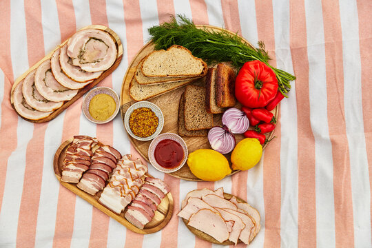 Top View of Outdoor Picnic Spread with Cold Cuts, Sliced Bread, Fresh Vegetables, and Sauces on Striped Blanket